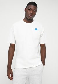 Mann trägt ein weißes T-Shirt mit kleinem blauen Logo auf der Brust und hellgraue Sweatpants, steht mit einer Hand in der Tasche vor einem weißen Hintergrund.