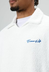 Witte pullover met textuur en kraag, met blauwe geborduurde tekst "Essence of Life" op de borst. Zacht, pluizig materiaal.