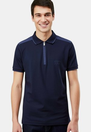 Jeune homme souriant portant un polo bleu marine à manches courtes avec une fermeture éclair et des panneaux d'épaule contrastants.