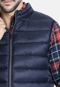 Chaleco acolchado azul marino con un alto cuello, presenta un cierre frontal y un logo redondo. Debajo, una camisa a cuadros en rojo, verde y azul marino.