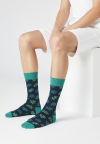 Osoba v bílých šortkách sedící na bílém bloku, obuv s námořnickými ponožkami se zelenými listovými vzory a tyrkysovými lemy a špičkami.