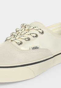 Off-white αθλητικό παπούτσι Vans με μαύρα και λευκά κορδόνια με σχέδιο, καπάκι από σουέντ στο μπροστινό μέρος και μαύρη ρίγα κατά μήκος της καουτσούκ σόλας.