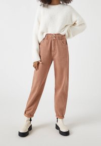 Kalhoty s vysokým pasem v terakotové barvě z bavlny, volného střihu, se dvěma předními kapsami, ohrnutými manžetami a zapínáním na knoflík. V kombinaci s bílým chlupatým svetrem.