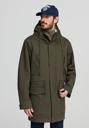 Giacca parka verde oliva con cappuccio, due grandi tasche frontali, coulisse e vestibilità relaxed, indossata sopra un dolcevita beige e jeans scuri.