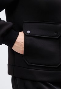 Χέρι μερικώς μέσα σε μαύρη τσέπη σακακιού με καπάκι που ασφαλίζει με δύο μαύρα κουμπιά με πατίνια, πάνω σε ανοιχτόχρωμο φόντο.