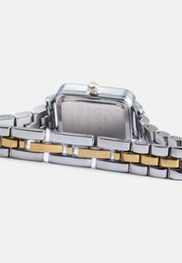 Reloj de metal en plata y oro con una cara cuadrada, correa de acero inoxidable que presenta elementos alternando brillantes en plata y oro, y una corona en el lateral.