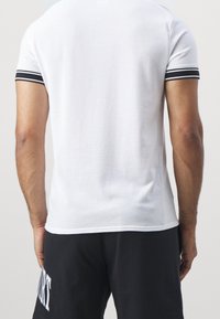 Maglietta sportiva bianca in rete, maniche corte con accenti a strisce nere, caratterizzata da una vestibilità rilassata e una texture traspirante. Indossata con pantaloni shorts neri.