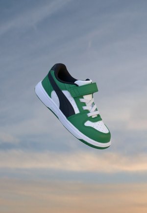 Baskets pour enfants vertes, blanches et noires avec lacets et bande velcro flottant sur un fond de ciel nuageux.