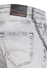 Jean en denim gris avec une étiquette en cuir, doté d'un détail en chaîne attaché à une boucle de ceinture et de coutures bien visibles sur les poches arrière.
