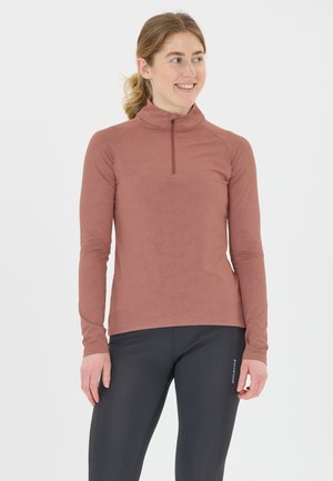 Kvinde der smiler og ser til højre, iført en rustfarvet langærmet top med en frontlynlås og sorte træningsleggings.