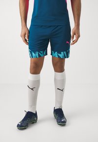 Marineblaue Sportshorts mit einem geometrischen türkisen Muster am Saum, kombiniert mit weißen Socken mit schwarzem Branding und schwarzen Fußballschuhen.