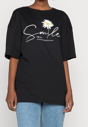 Personne portant un T-shirt noir avec le texte blanc en cursive « Smile » et un motif de marguerite, associé à un jean en denim bleu clair.
