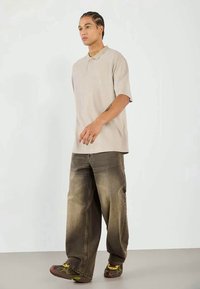 Polo bege de mangas curtas, combinado com jeans castanhos desbotados e oversized, e ténis com padrão. Design simples, corte descontraído e estilo casual.