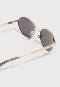 Kerek napszemüveg sötét lencsékkel, arany fém kerettel, átlátszó orrtámaszokkal és texturált arany részletekkel a szárakon.