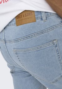 Vaaleansiniset farkut, joissa on teksturoitu pinta, taka-taskut ja ruskea nahkalappu, jossa lukee "ONLY & SONS PASSIONATE JEANS MAKERS."