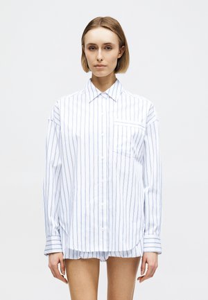 Młoda kobieta z krótkimi blond włosami, ubraną w dużą białą koszulę zapinaną na guziki z cienkimi niebieskimi pionowymi paskami oraz pasujące szorty, na jednolitym tle.