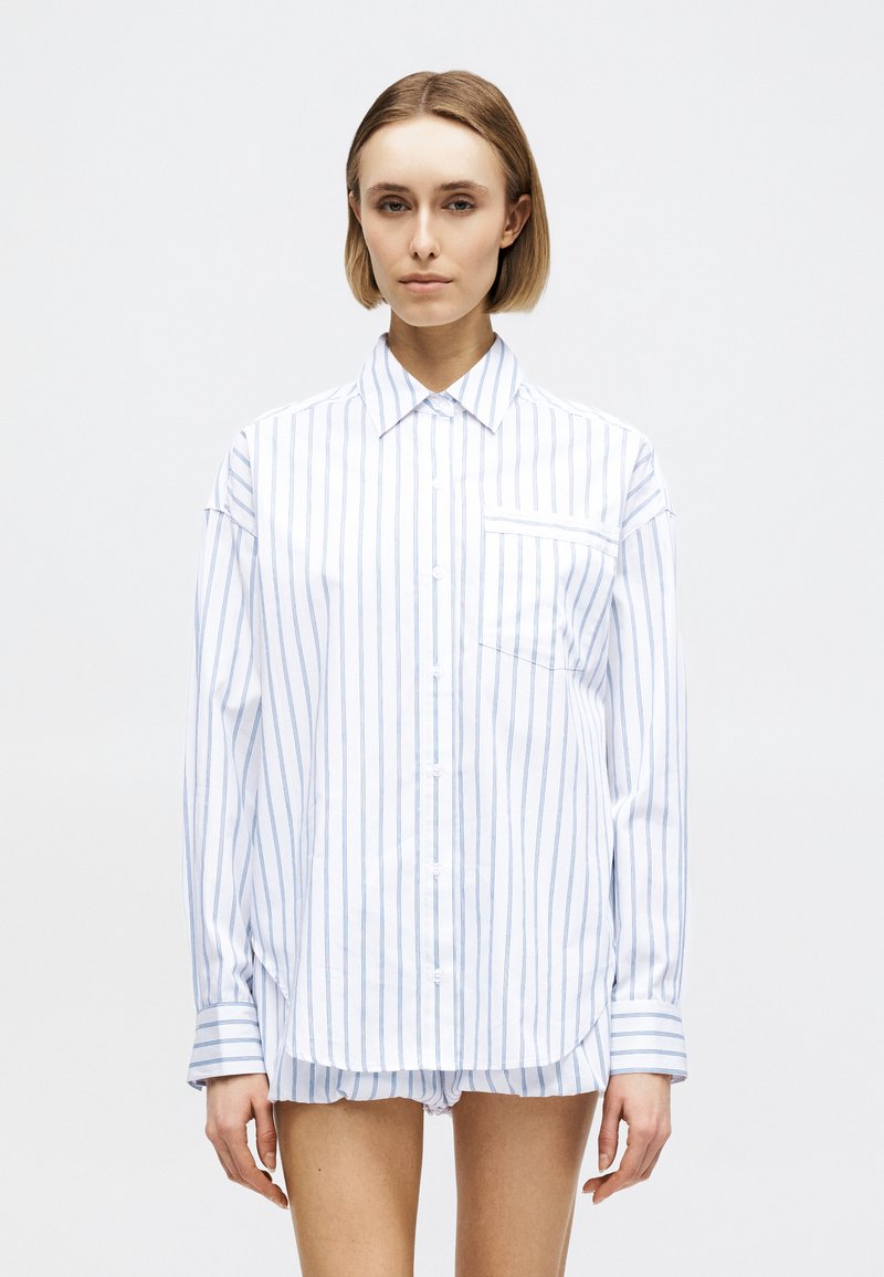 Νεαρή γυναίκα με κοντά ξανθά μαλλιά που φοράει υπερμεγέθη λευκό πουκάμισο με λεπτές μπλε κάθετες ρίγες και ταιριαστό σορτς, σε απλό φόντο.