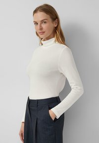 White turtleneck top with long sleeves, smooth fabric, form-fitting design, paired with dark tailored trousers featuring a pocket.