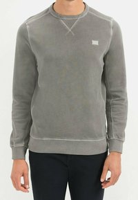Grauer Pullover aus weichem Stoff. Mit gerippten Manschetten und Saum, Rundhalsausschnitt und einem kleinen Logo-Patch auf der Brust. Glatte Textur.