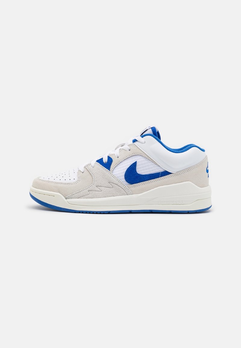 Basket basse en daim blanc et beige avec des accents bleus, arborant un swoosh Nike bleu et des lacets blancs sur un fond neutre.