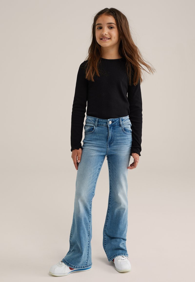 Fille debout portant un haut à manches longues noir, un jean flare bleu clair et des baskets blanches sur un fond beige uni.