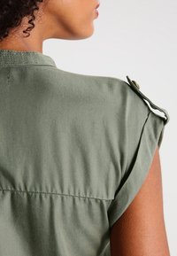 Zbliżenie na ramię osoby noszącej oliwkowozieloną koszulkę bez rękawów z zapinanym na guzik paskiem na ramieniu, widziane od tyłu.