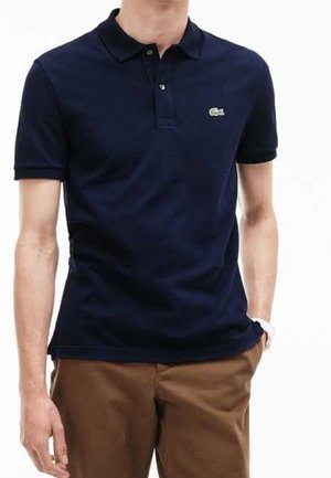 Homme portant un polo bleu marine à manches courtes avec col et un pantalon marron, debout, une main dans la poche.