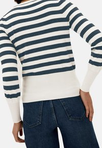 Maglione a righe navy e bianco, con polsini e orlo a coste, abbinato a jeans in denim scuro. Il tessuto sembra morbido e ha un fit casual.
