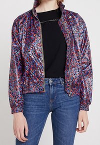 Veste bomber colorée et à motifs avec une finition brillante, présentant une fermeture éclair à l'avant, des poignets en côtes et une ceinture en côtes. Portée par-dessus une chemise noire.