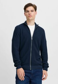 Sötétkék, cipzáros pulóver texturált mintázattal, alatta fehér pólóval és bordázott mandzsettákkal valamint rózsás aljjal. A modell egy sima háttér előtt pózol.