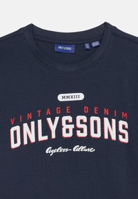 T-shirt bleu marine avec "ONLY & SONS" en grandes lettres blanches et rouges, "VINTAGE DENIM" au-dessus, et "Ageless Allure" en dessous en écriture cursive.