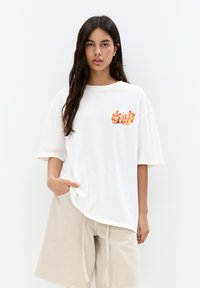 T-shirt bianco oversize con un design grafico colorato che cola sul lato sinistro del petto, abbinato a pantaloni corti beige con coulisse.