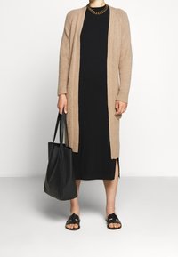 Beżowy dziany kardigan noszony na czarnej sukience, zestawiony z czarnymi sandałami i dużą czarną torbą na ramię. Posiada otwarty przód i długie rękawy.