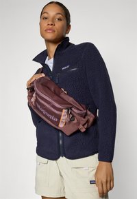 Giacca in pile blu navy con tasche con zip, abbinata a una borsa a tracolla bordeaux con un accento arancione e il logo "Patagonia", indossata a tracolla.