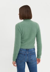 Maglione verde a collo alto, design corto, texture morbida, indossato con jeans blu a vita alta, sfondo semplice, vista posteriore.