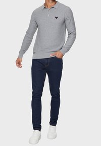 Hombre con camiseta de polo de manga larga texturizada en gris claro, jeans ajustados oscuros y zapatillas blancas, de pie con una mano en el bolsillo.