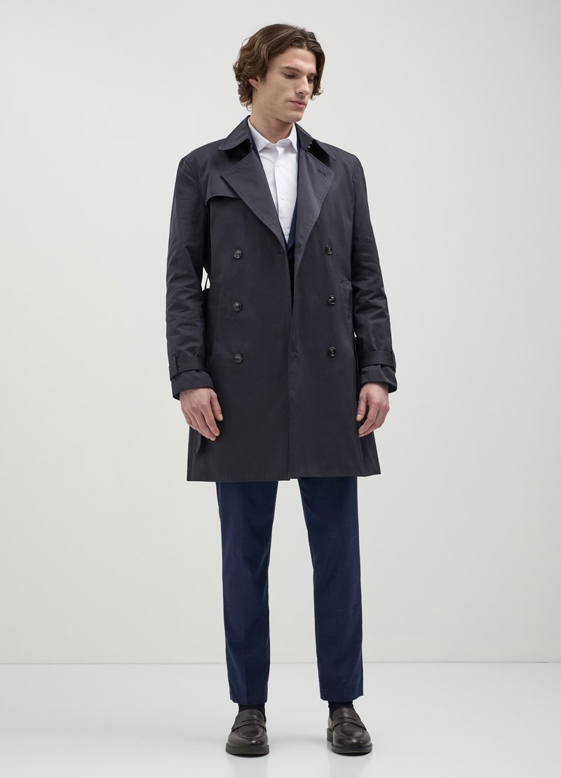Manteau trench double boutonnage en tissu bleu marine avec un col structuré, des accents de boutons et une coupe ajustée, associé à un pantalon bleu marine et des chaussures.