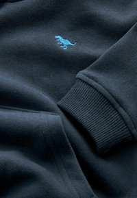 Sudadera azul marino oscura con puños y dobladillo acanalados, que presenta un pequeño bordado de un dinosaurio turquesa en el pecho. Textura de tela suave.