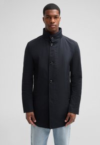 Navy blue coat featuring a high collar, button front, and side pockets. Made from a smooth fabric with a tailored fit.