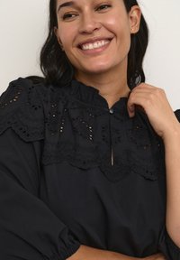 Blusa negra con cuello mandarín, que presenta intrincados bordados de encaje en los hombros y las mangas. Tejido texturizado con detalles fruncidos.