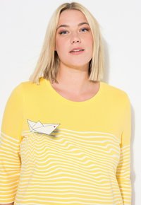 Gul långärmad skjorta med vita vågiga ränder och en pappersbåt med ett rött hjärta. Mjuk tyg med rundad halslinje.