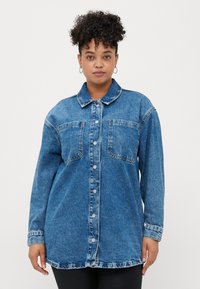 Woman with curly hair wearing oversized blue denim jacket with front pockets and silver buttons, standing against plain white background.
