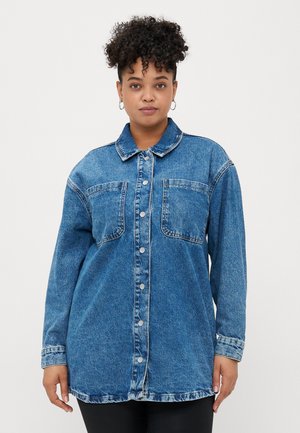 Woman with curly hair wearing oversized blue denim jacket with front pockets and silver buttons, standing against plain white background.