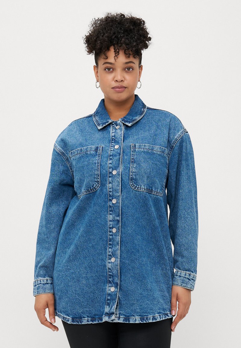 Femme aux cheveux bouclés portant une grande veste en jean bleu avec des poches avant et des boutons en argent, se tenant devant un fond blanc uni.