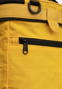 Zaino in tessuto giallo con superficie testurizzata, dotato di una tasca con zip nera e accessori neri. Chiusura con un linguetta rotonda.