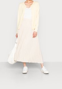 Kobieta ubrana w jasnżółty kardigan, biały top, beżową plisowaną spódnicę, białe trampki, trzymająca białą torbę na jednolitym tle.