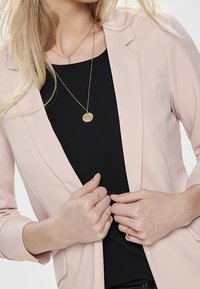 Light pink blazer made of smooth fabric with a tailored design. Worn over a black top, featuring layered gold necklaces and rings.