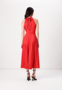 Robe rouge à col halter avec un corsage ajusté, une silhouette à jambes larges, un dos nu et un détail de nœud au cou. Portée avec des talons noirs à lanières.