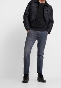 Man som bär svart jacka, svart tröja, urblekta grå jeans med uppvikta ben och svarta högskor som står med händerna i fickorna.