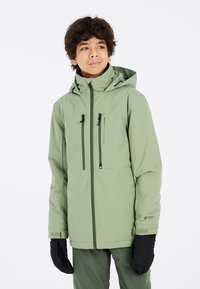 Giacca impermeabile verde con cappuccio, zip frontale, due tasche sul petto e polsini elasticizzati. Indossata con guanti neri e pantaloni verdi coordinati.